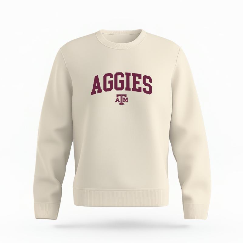 The Ivory Unisex Aggies Varsity Organic Cotton Crewneck is set against a Ghost - White Studio background. The Aggies Varsity graphic is in a Sports Graphics style.