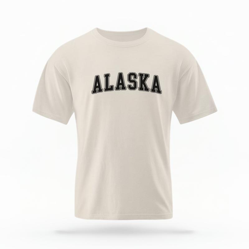 The Ivory Unisex Alaska Varsity T-shirt is set against a Ghost - White Studio background. The Alaska Varsity graphic is in a Varsity style.
