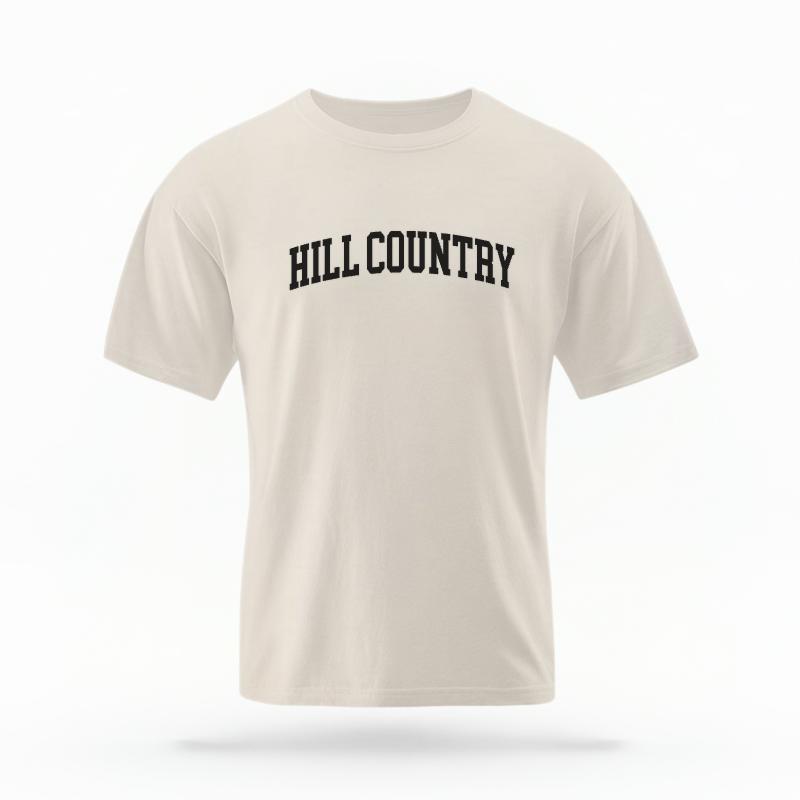 The Ivory Unisex Hill Country Varsity T-shirt is set against a Ghost - White Studio background. The Hill Country Varsity graphic is in a Varsity style.