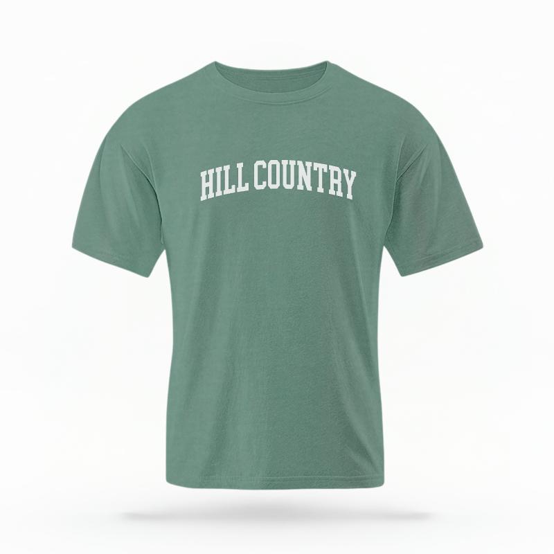 The Light Green Unisex Hill Country Varsity T-shirt is set against a Ghost - White Studio background. The Hill Country Varsity graphic is in a Varsity style.