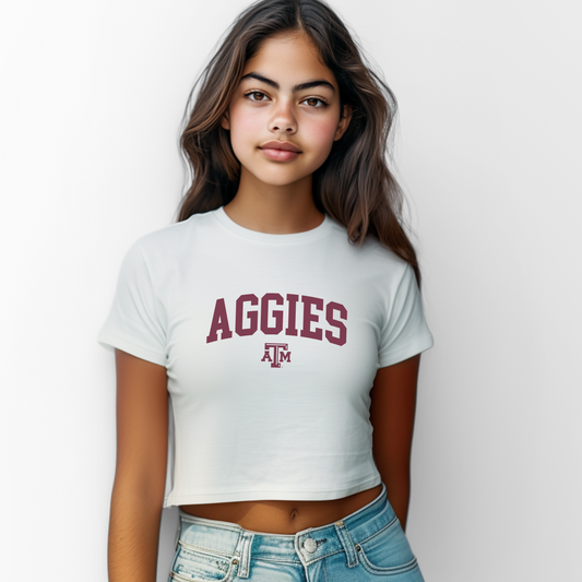 The White Female Aggies Varsity Women's Baby Tee is set against a Ghost - White Studio background. The Aggies Varsity graphic is in a Varsity style.