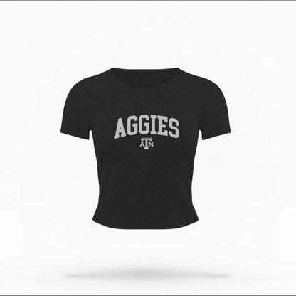The Black Female Aggies Varsity Women's Baby Tee is set against a Ghost - White Studio background.  The Aggies Varsity graphic is in a Varsity style. 