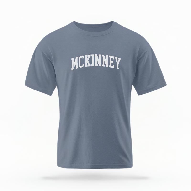The Blue Jean Unisex McKinney Varsity T-shirt is set against a Ghost - White Studio background.  The McKinney Varsity graphic is in a Varsity style. 