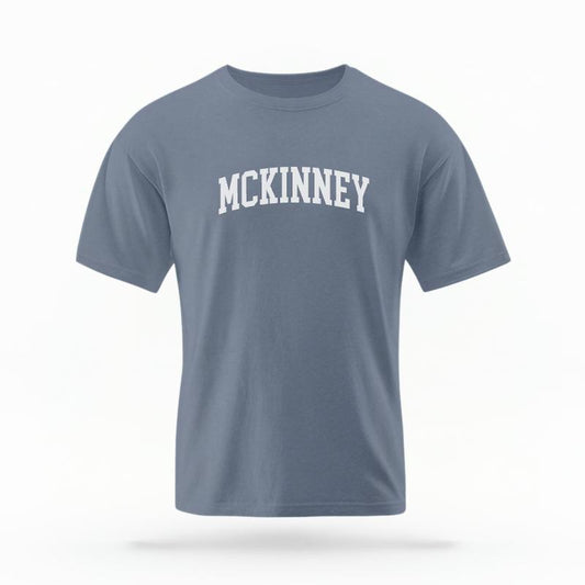 The Blue Jean Unisex McKinney Varsity T-shirt is set against a Ghost - White Studio background.  The McKinney Varsity graphic is in a Varsity style. 