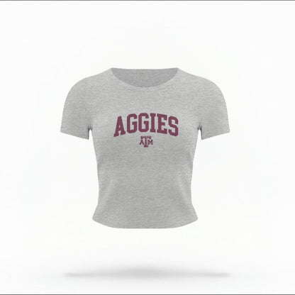 The Heather Grey Female Aggies Varsity Women's Baby Tee is set against a Ghost - White Studio background.  The Aggies Varsity graphic is in a Varsity style. 