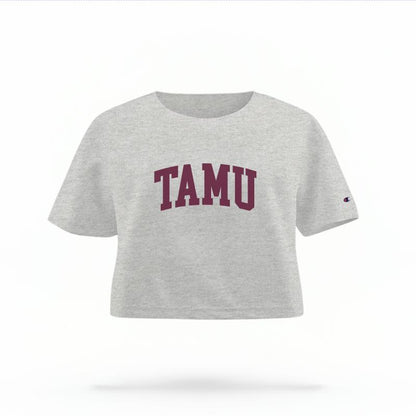 The Heather Grey Female TAMU Varsity Women's Boxy Crop Top is set against a Ghost - White Studio background.  The TAMU Varsity graphic is in a Sports Graphics style. 