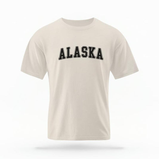 The Ivory Unisex Alaska Varsity T-shirt is set against a Ghost - White Studio background.  The Alaska Varsity graphic is in a Varsity style. 