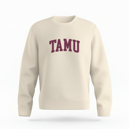 The Ivory Unisex TAMU Varsity Organic Cotton Crewneck is set against a Ghost - White Studio background.  The TAMU Varsity graphic is in a Sports Graphics style. 