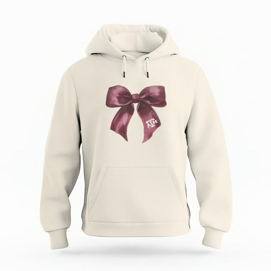 The Ivory Unisex Texas A&M Coquette Bow Organic Cotton Hoodie is set against a Ghost - White Studio background.  The Texas A&M Coquette Bow graphic is in a Coquette style. 