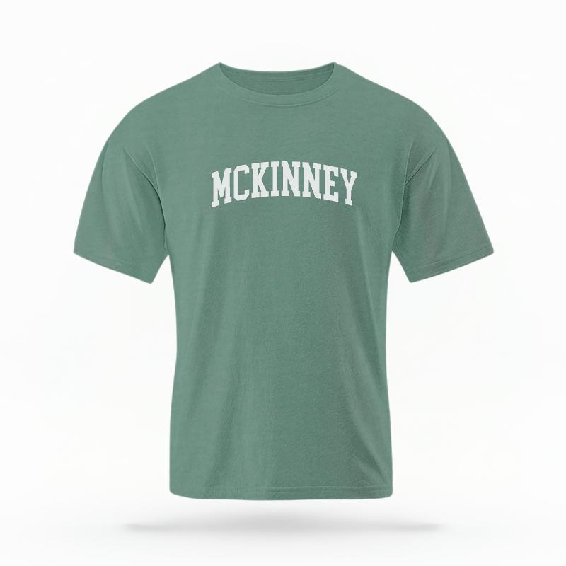The Light Green Unisex McKinney Varsity T-shirt is set against a Ghost - White Studio background.  The McKinney Varsity graphic is in a Varsity style. 