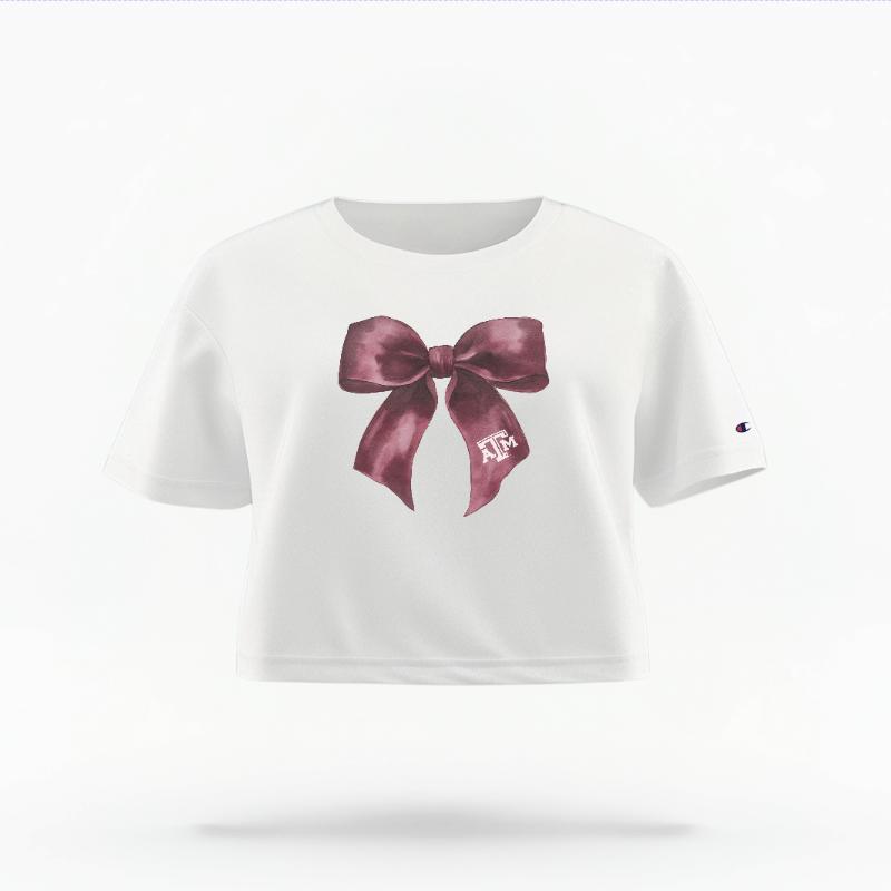 The White Female Texas A&M Coquette Bow Women's Boxy Crop Top is set against a Ghost - White Studio background.  The Texas A&M Coquette Bow graphic is in a Coquette style. 