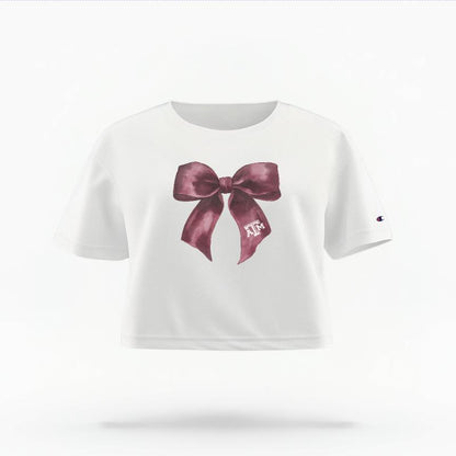 The White Female Texas A&M Coquette Bow Women's Boxy Crop Top is set against a Ghost - White Studio background.  The Texas A&M Coquette Bow graphic is in a Coquette style. 