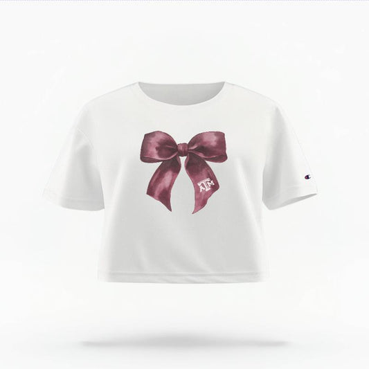 The White Female Texas A&M Coquette Bow Women's Boxy Crop Top is set against a Ghost - White Studio background.  The Texas A&M Coquette Bow graphic is in a Coquette style. 